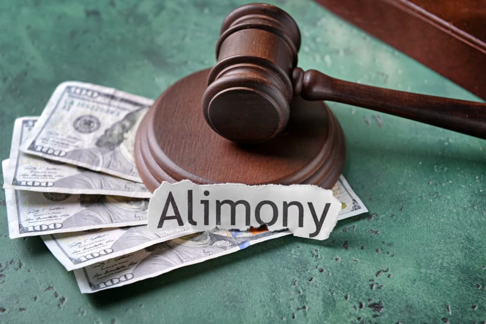 A judge's gavel, stacks of money, and a document labeled "ALIMONY" lying on a table.






