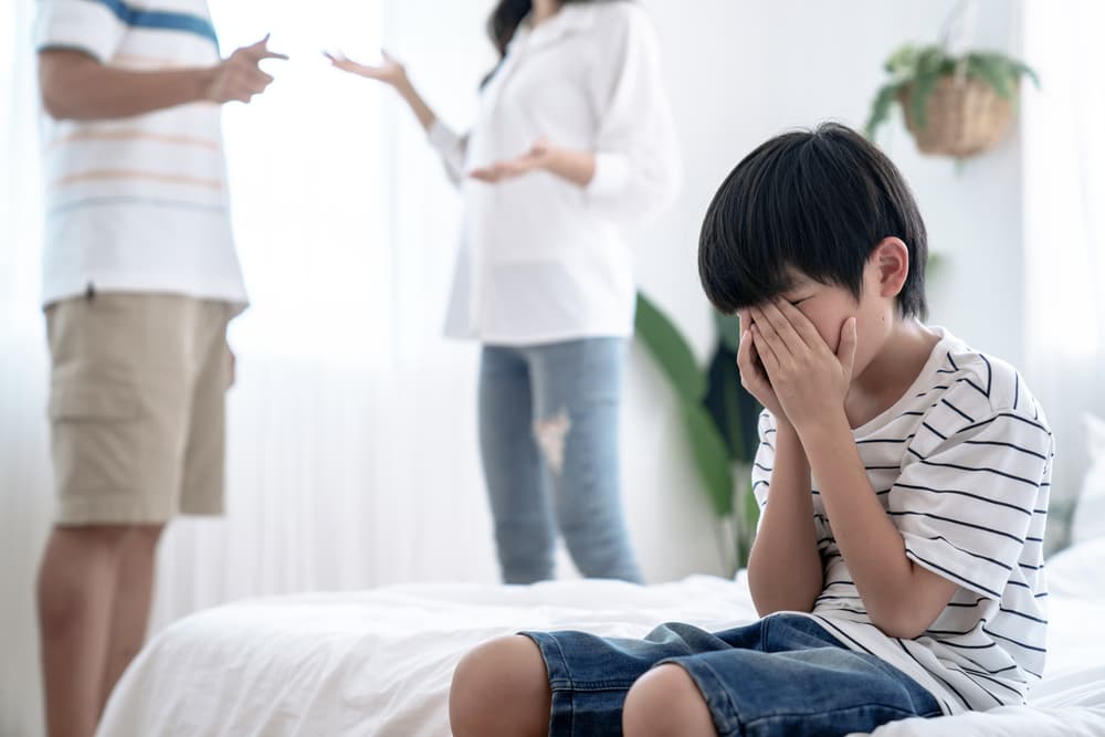 A crying little Asian boy sits in the foreground, distressed, while his parents argue in the background, highlighting family problems and divorce struggles.