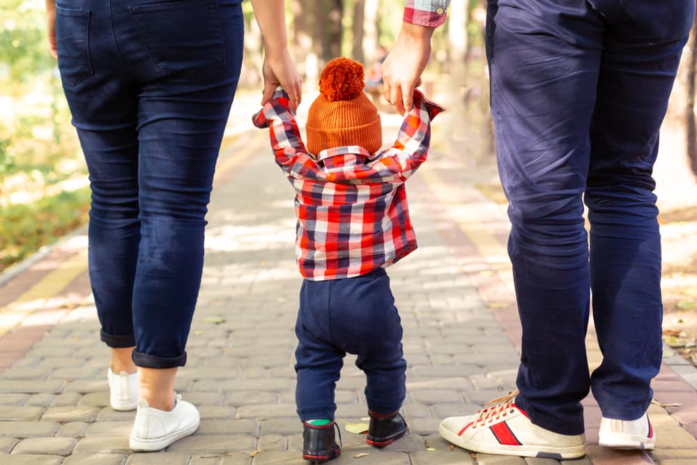 Mom and Dad walk through the park, holding their young son by the arms as they lead him along.