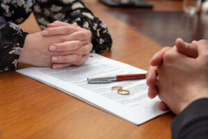 Signing divorce and marriage dissolution documents. Husband and wife’s hands, wedding rings visible, with legal papers on a wooden surface awaiting signatures.