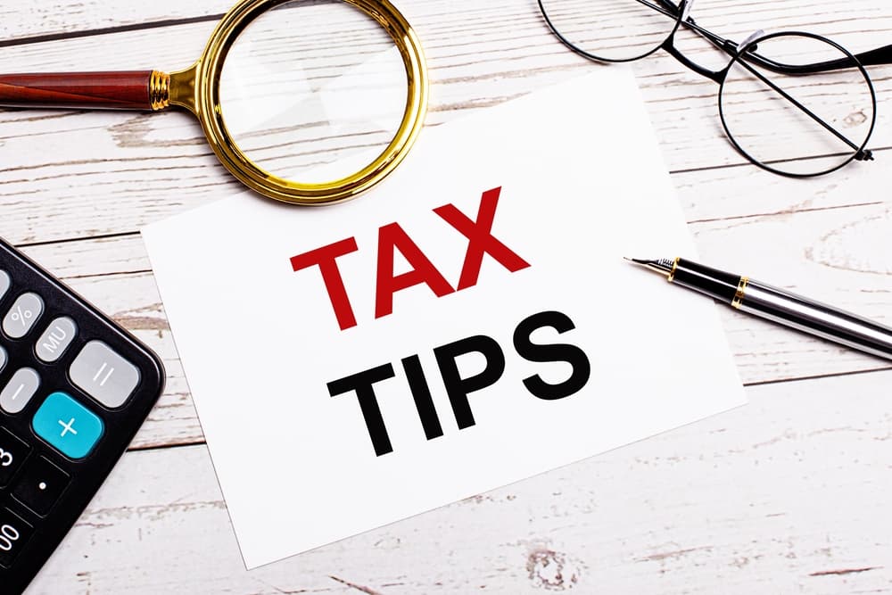A workplace with a pen, glasses, a calculator, a magnifying glass and a white sheet of paper with the text of the TAX TIPS on a white wooden table